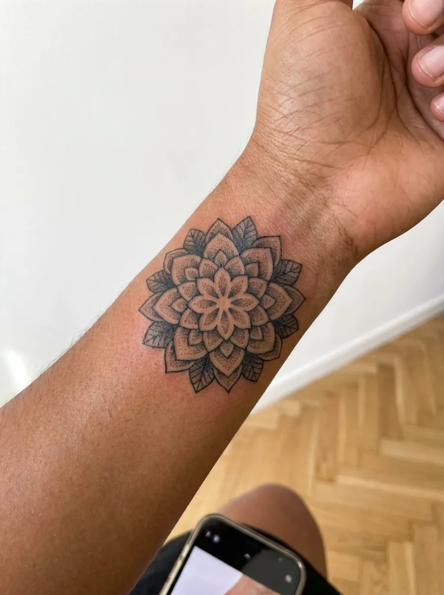 Petit tatouage dotwork circulaire au poignet sur peau mate, mandala floral en pétales superposés et ombrages pointillés noirs, photo naturelle lumière